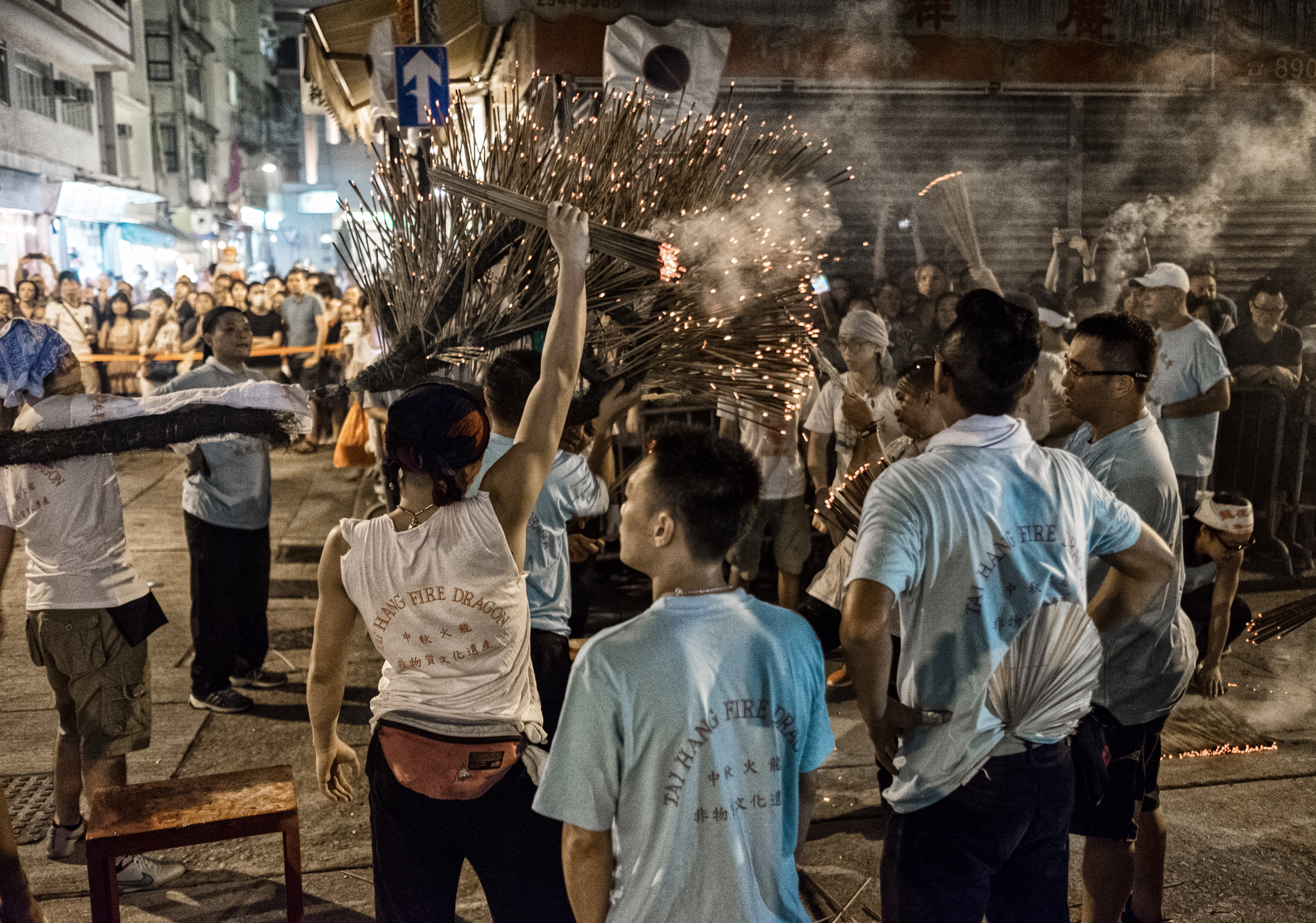 Crowd gathered at night as performers swing bundles of burning incense sticks, sending sparks into the air during a street ritual.