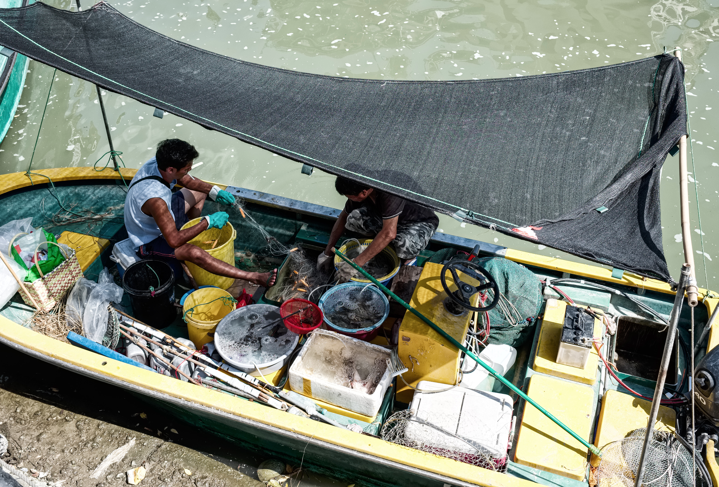 Two fishermen seated in a small boat, sorting nets and buckets under a stretched canopy beside green harbor water.