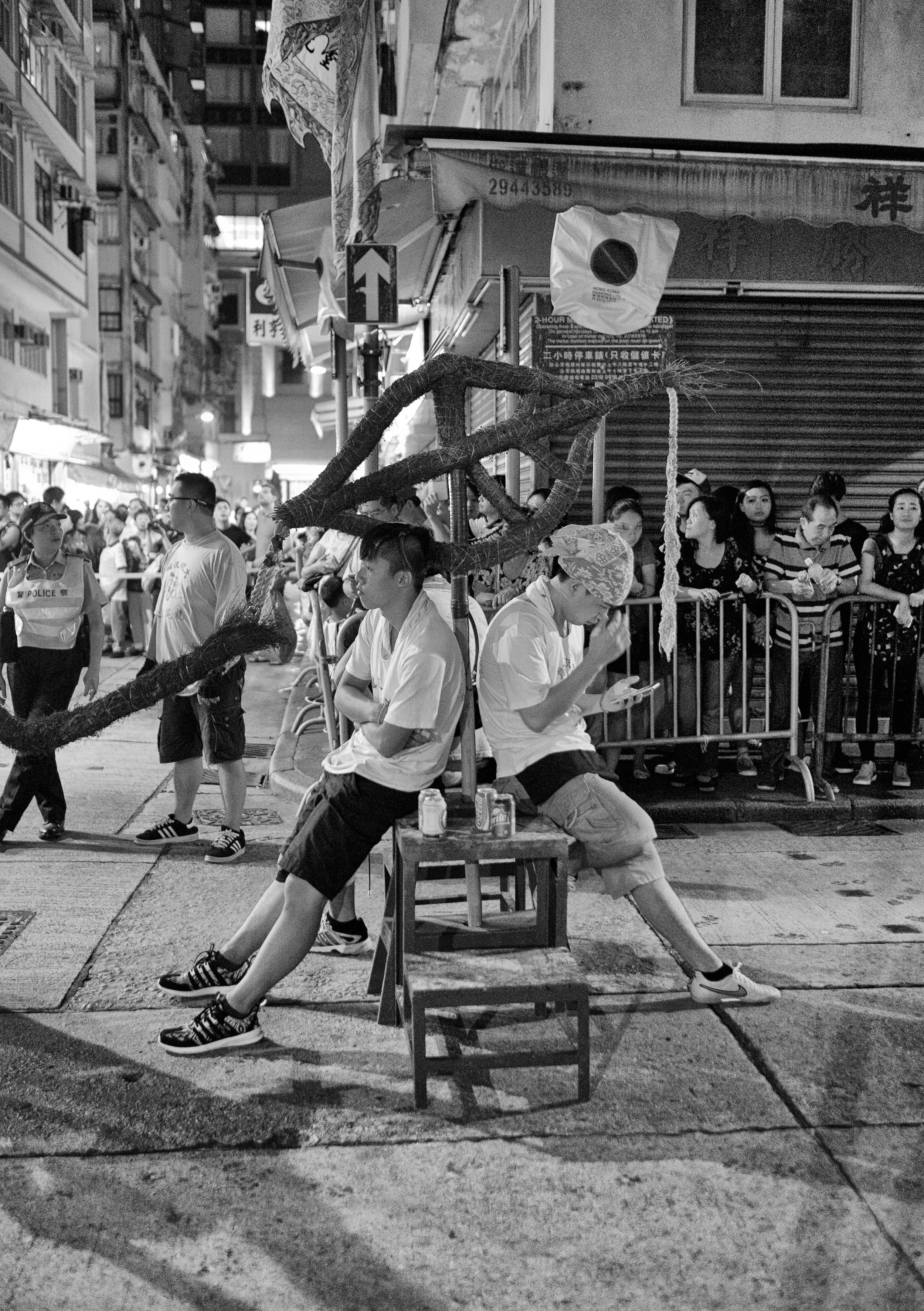 Two men resting beneath a festival structure on a crowded street at night