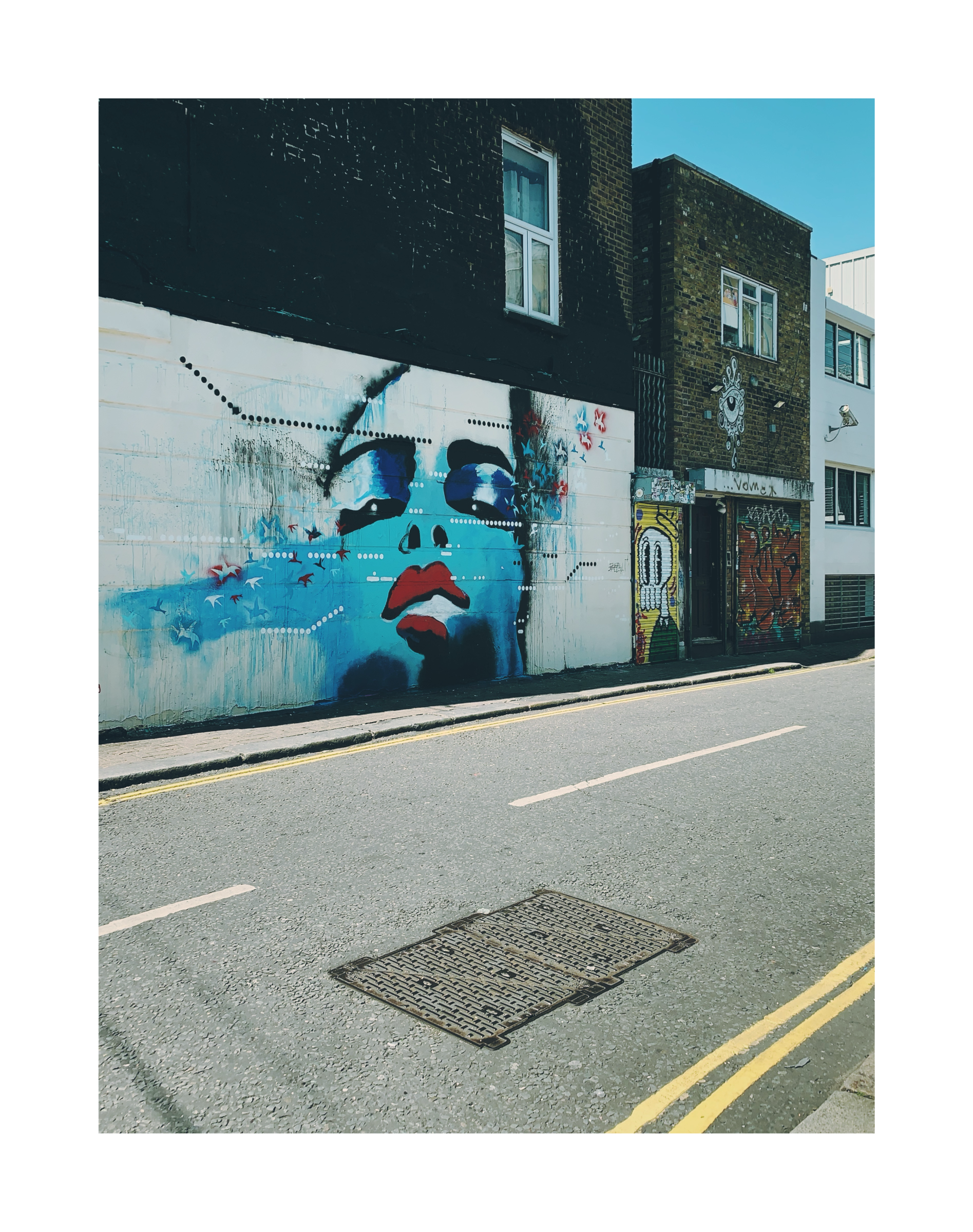 Street mural depicting a stylized blue face painted across a building wall, with surrounding urban textures and storefronts.